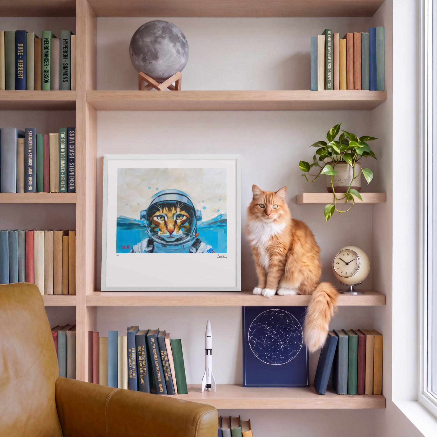 Cat Astronaut print in a white frame on an oak bookshelf, with Squash, the ginger cat it portrays, sitting beside it