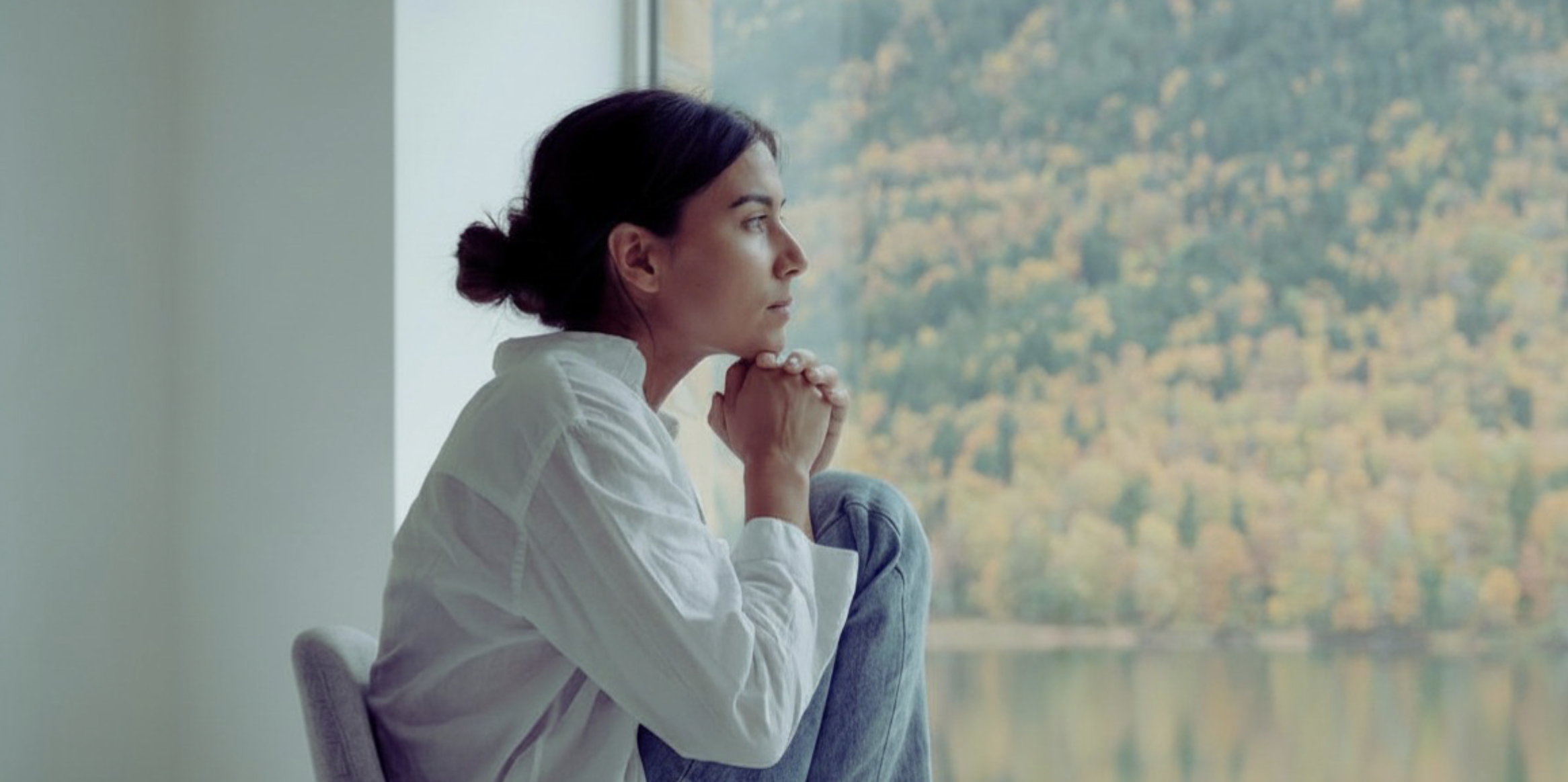 Young woman seated, looking out a window at a landscape of trees and a lake.