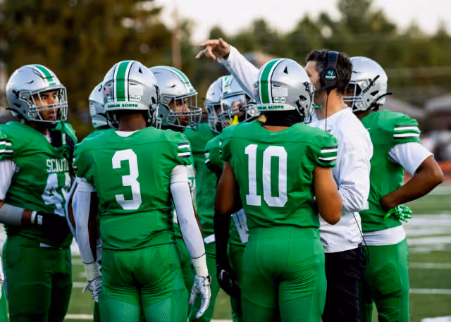 Football players in green uniforms and helmets huddle around a coach giving instructions on the field.