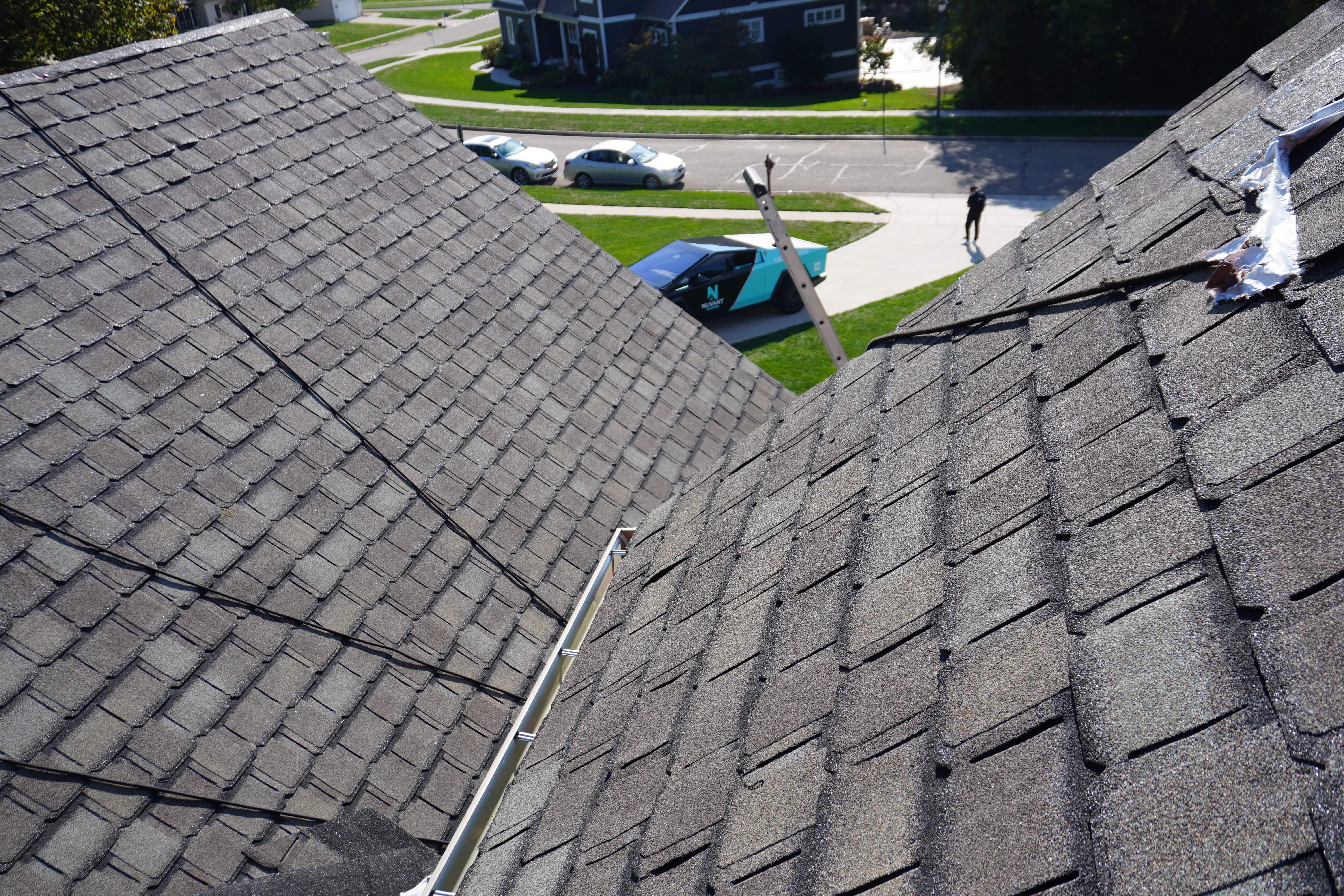 View of two gray shingled rooftops with a ladder against one roof and a parked teal and black truck on the street below.