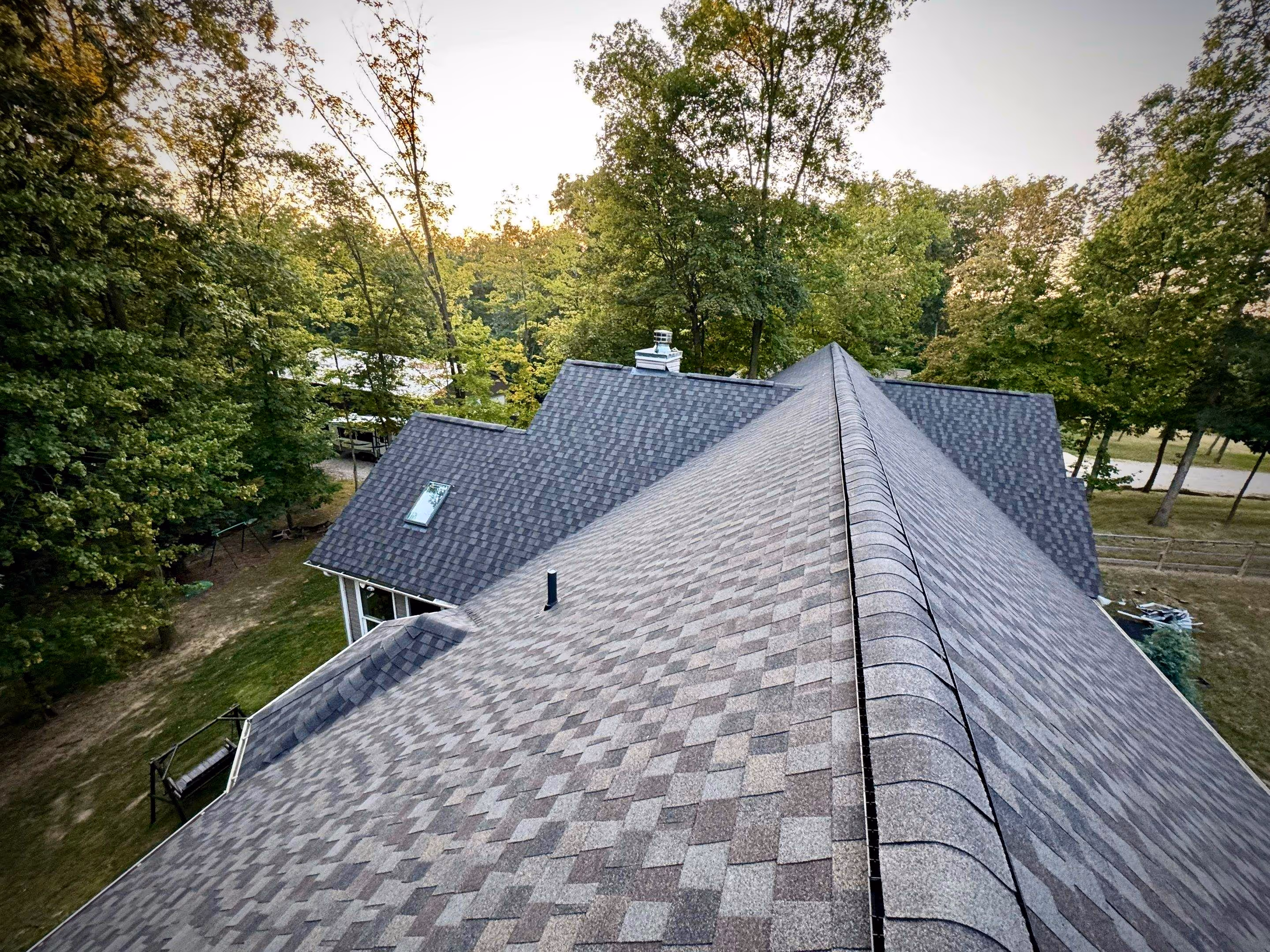 Aerial view of a house roof with gray asphalt shingles surrounded by green trees at sunset.