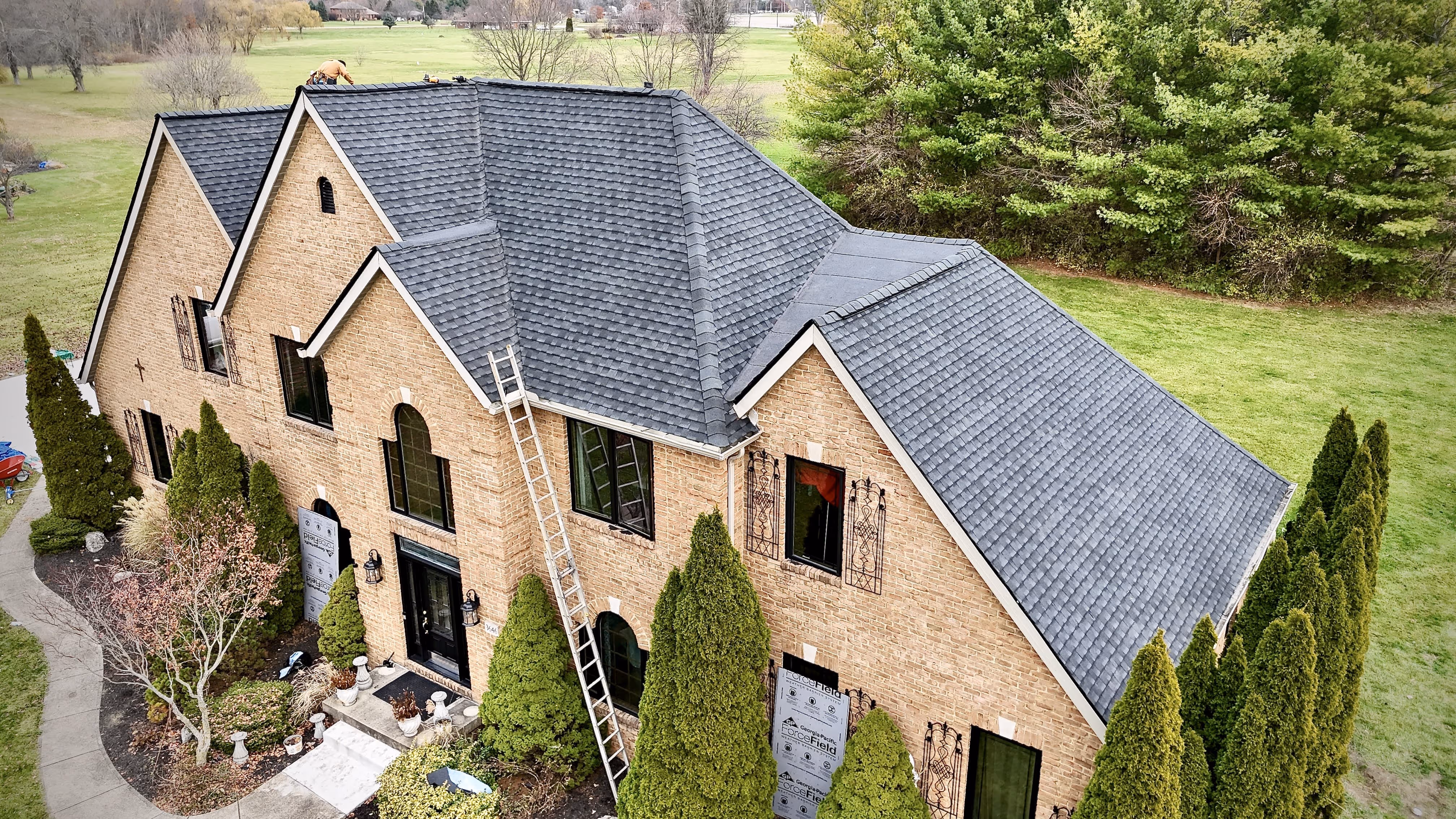 A large brick house with a newly installed gray shingle roof and a ladder leaning against it, surrounded by evergreen trees and a lawn.