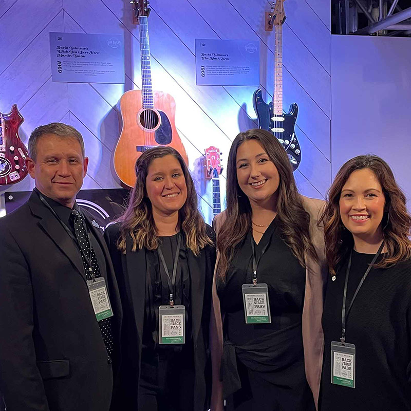 Three women and a man dressed in black with badges around their necks, at an event with guitars on the wall behind them.