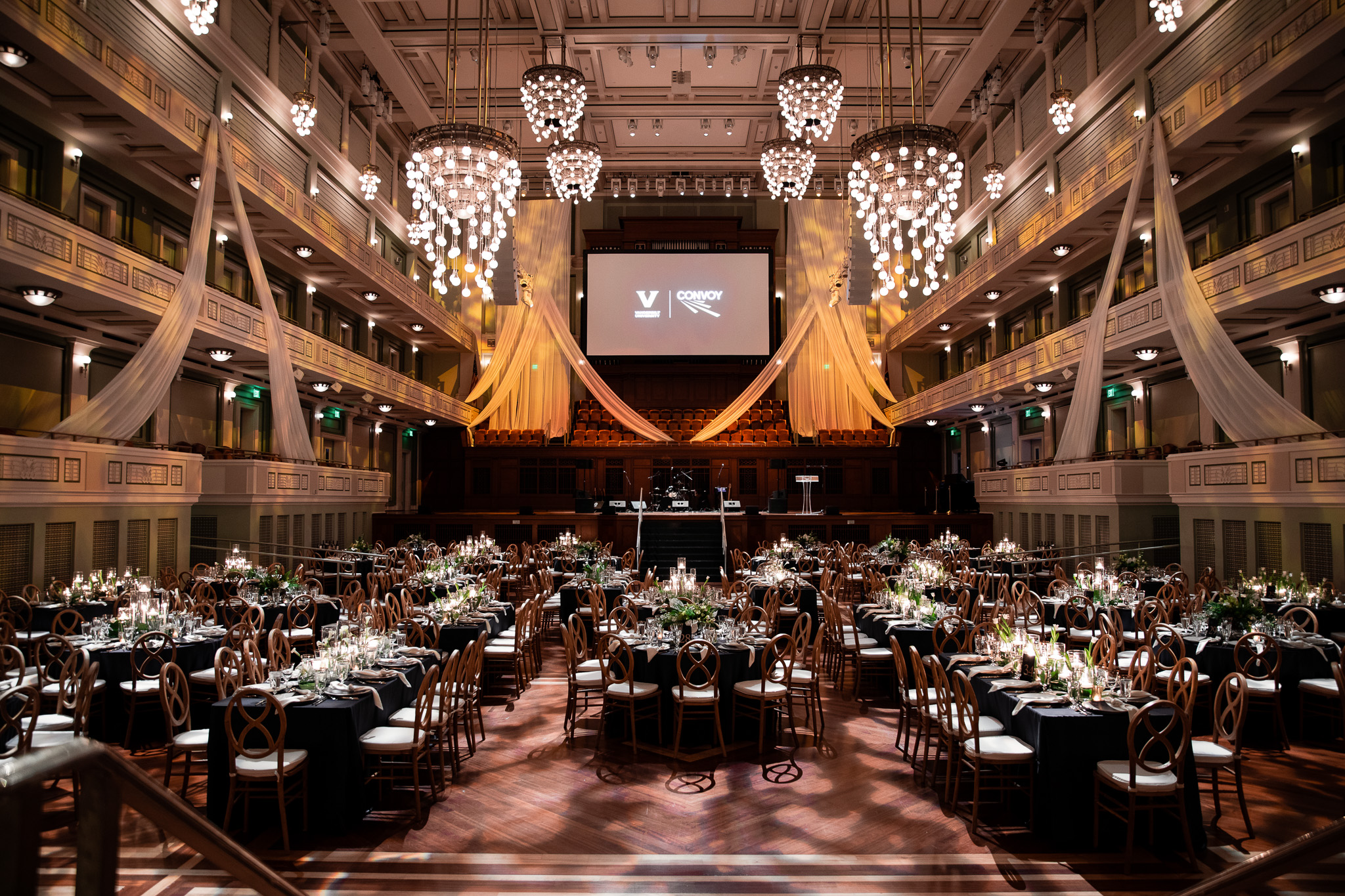 Elegant gala setup inside the Schermerhorn Symphony Center for the Convoy event, featuring round candlelit tables, draped fabric decor, and a stage with branded screens under grand chandeliers.