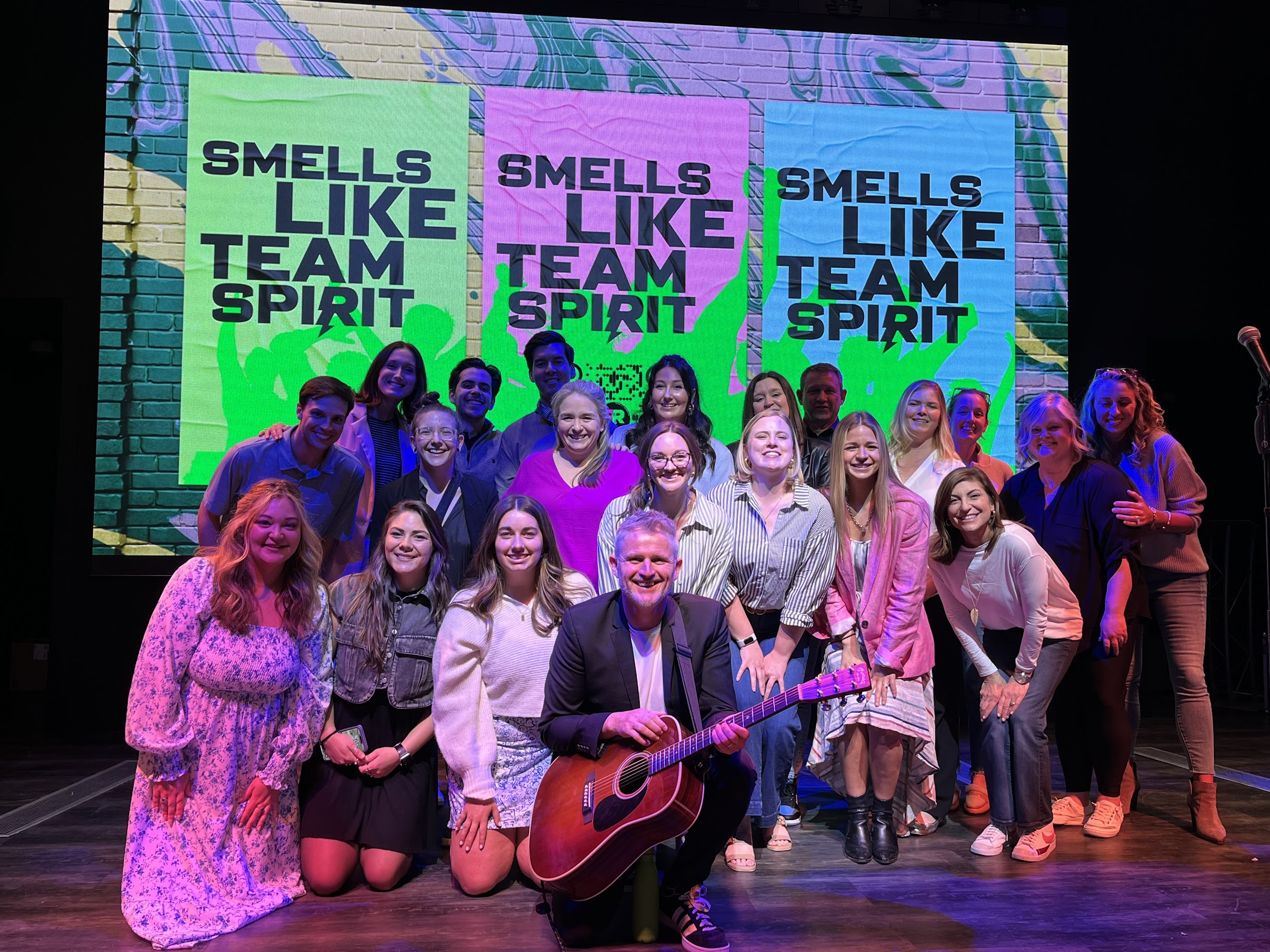 Group of people posing for a picture with a man holding a guitar in the front, and signs in behind them that say "Smells Like Team Spirit".