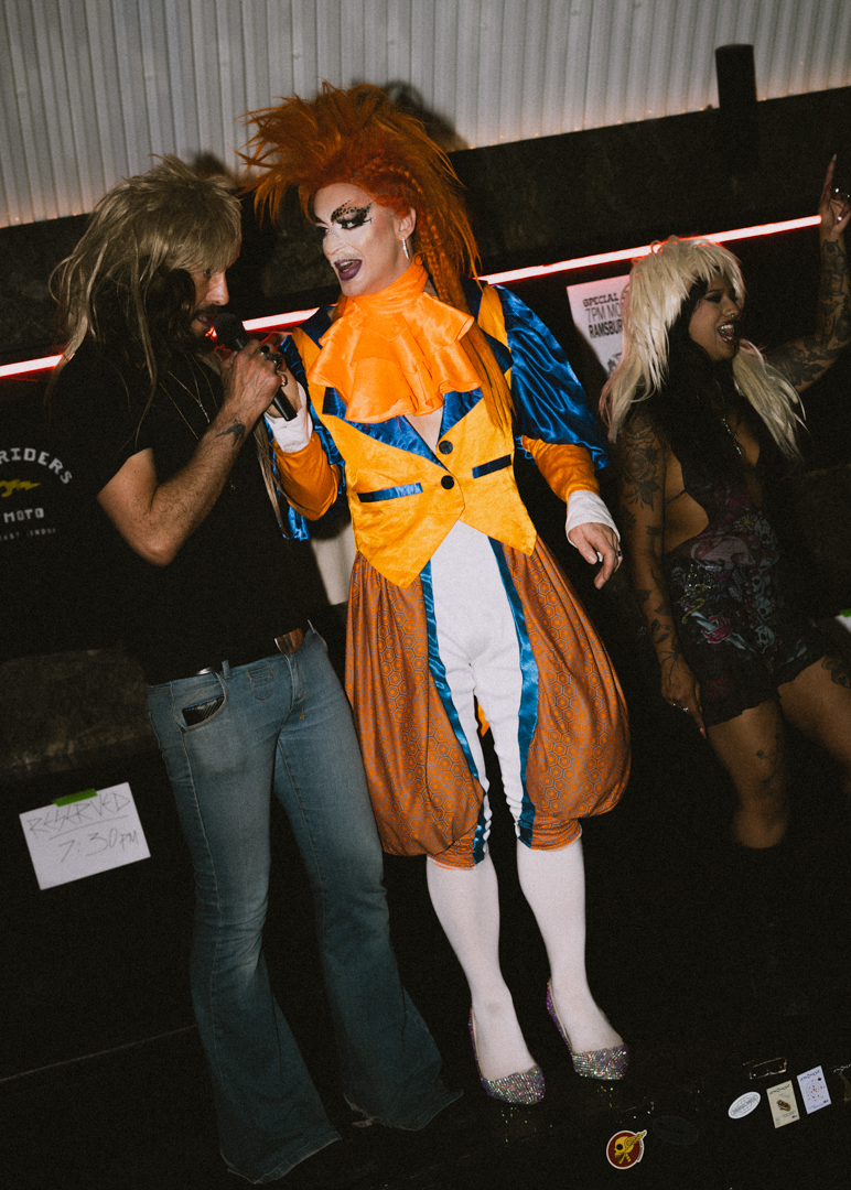 Three people dancing: a drag artist in orange and blue costume with bright makeup, another in casual clothes.