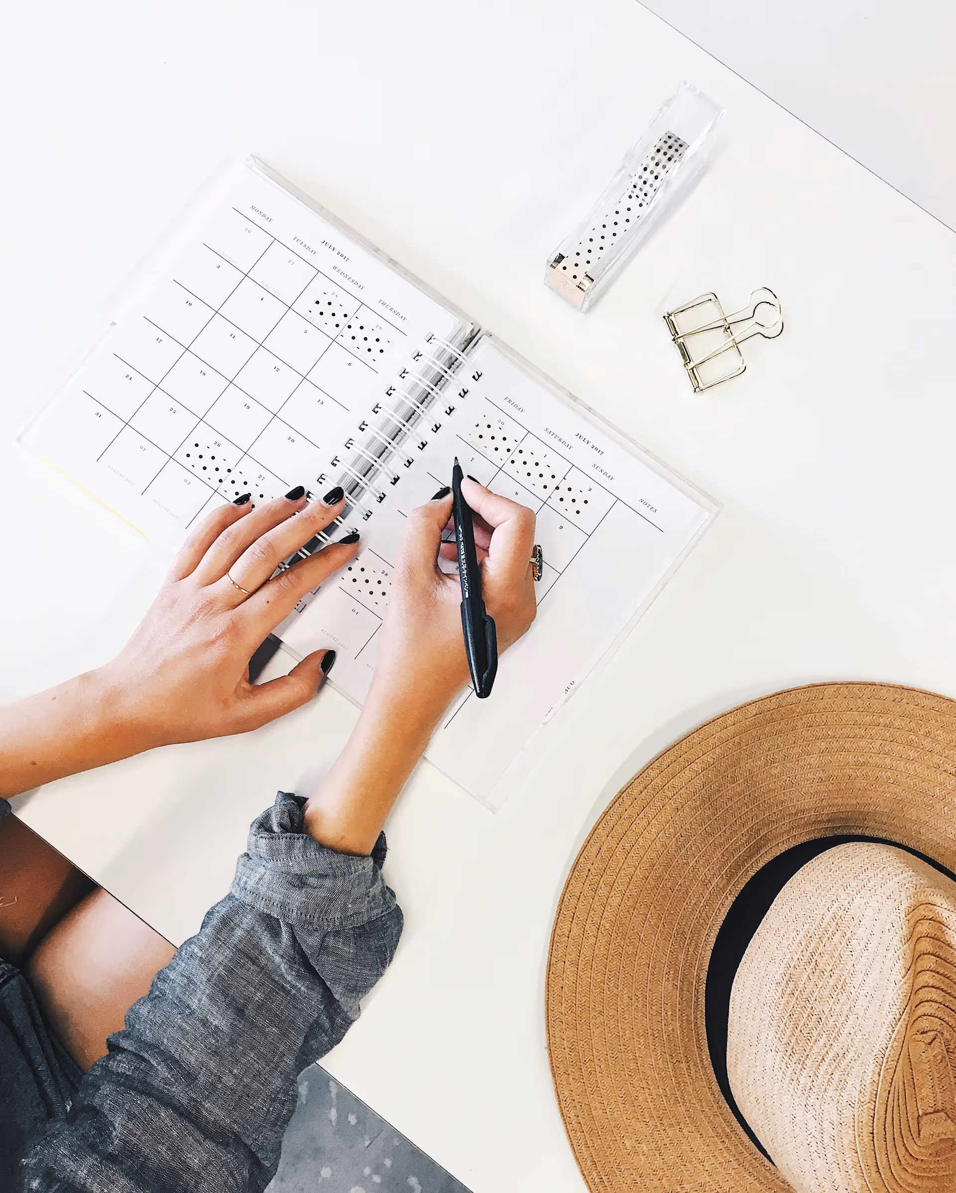 Woman writing in a notebook next to which is a hat. 