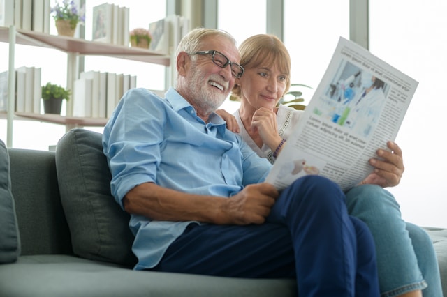 Elderly couple reading newspaper together on comfortable couch at home