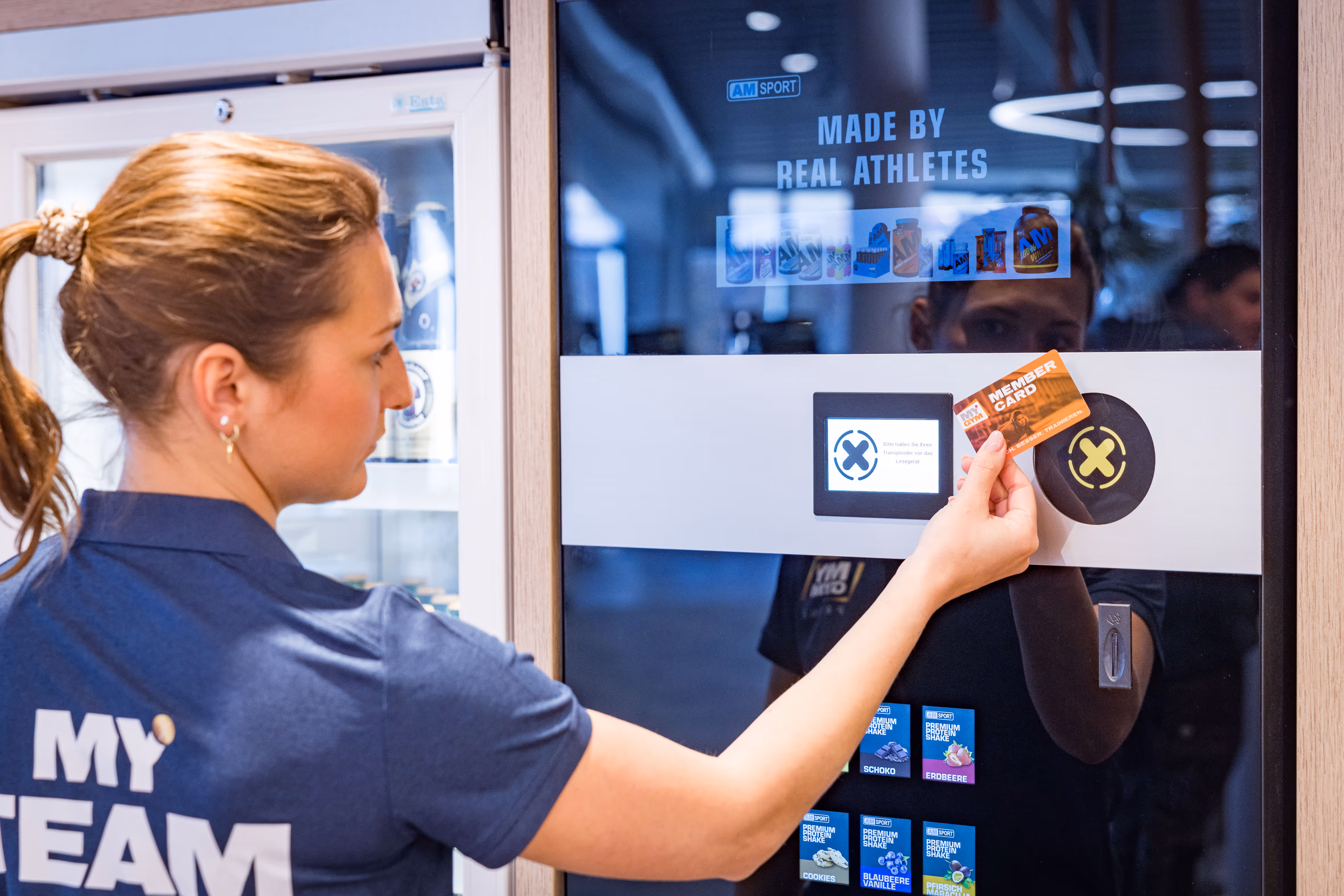 Frau scannt Mitgliedskarte an einem Automaten mit Text 'Made by Real Athletes' und Produktbildern.