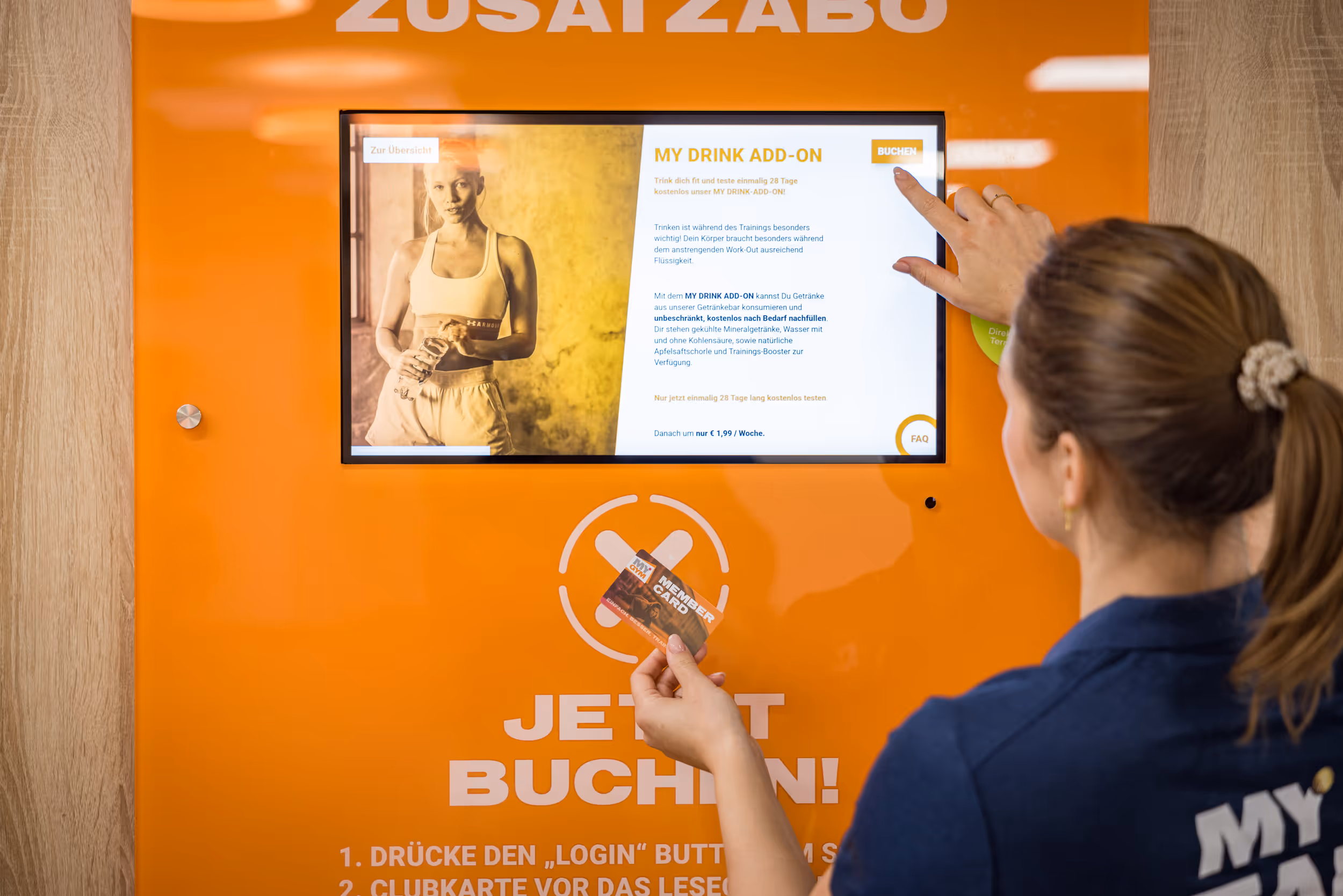 Frau bedient an orangefarbenem Bildschirmterminal, hält Mitgliedskarte und tippt auf ‚Buchen‘-Taste.