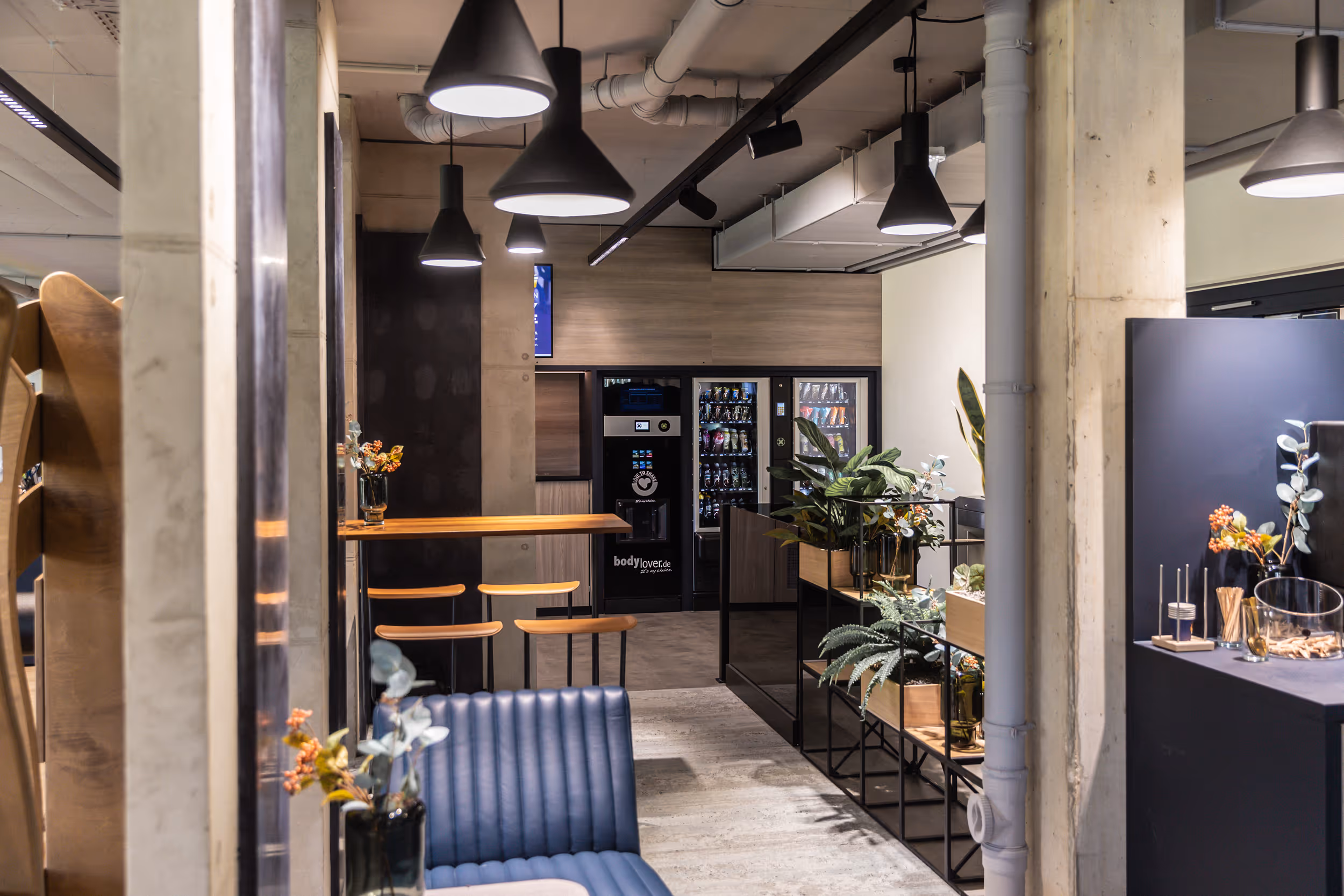 Moderner Pausenraum mit blauen Sitzbänken, hölzernen Barhockern, schwarzen Hängeleuchten und Snackautomaten im Hintergrund.