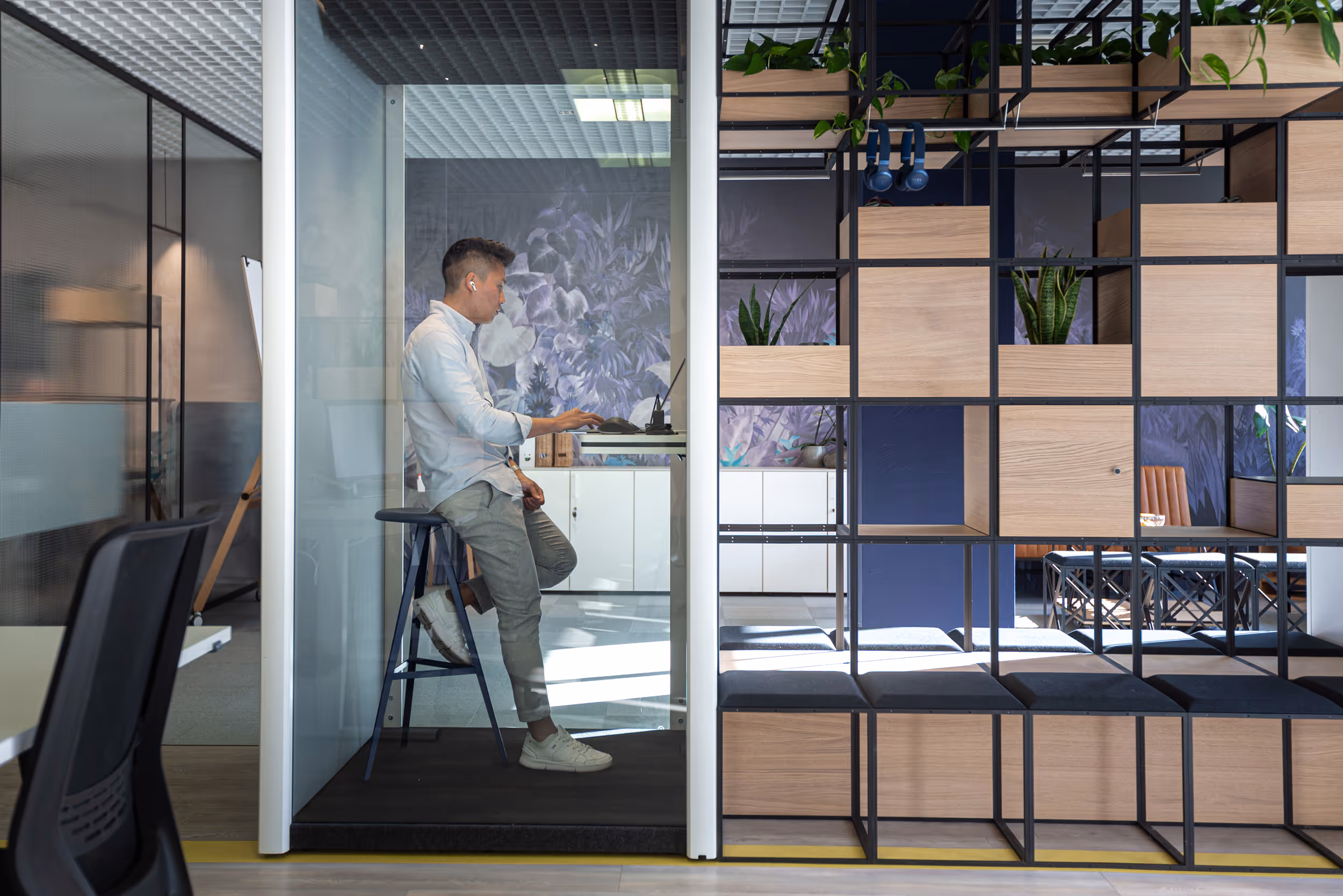 Ein Mann sitzt in einem gläsernen Arbeitsbereich auf einem Hocker und arbeitet an einem Laptop, neben modularen Holzregalen mit Pflanzen.