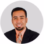 Portrait of a smiling man with a goatee wearing a black jacket and patterned orange tie against a light gray background.