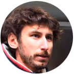 Close-up of a man with dark wavy hair, beard, and serious expression wearing a jacket.