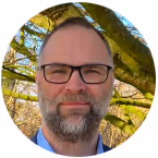 Man with short hair, beard, and glasses smiling outdoors with green tree branches in the background.