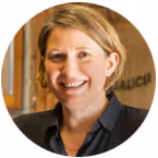 Smiling woman with short blonde hair wearing a dark blazer indoors.