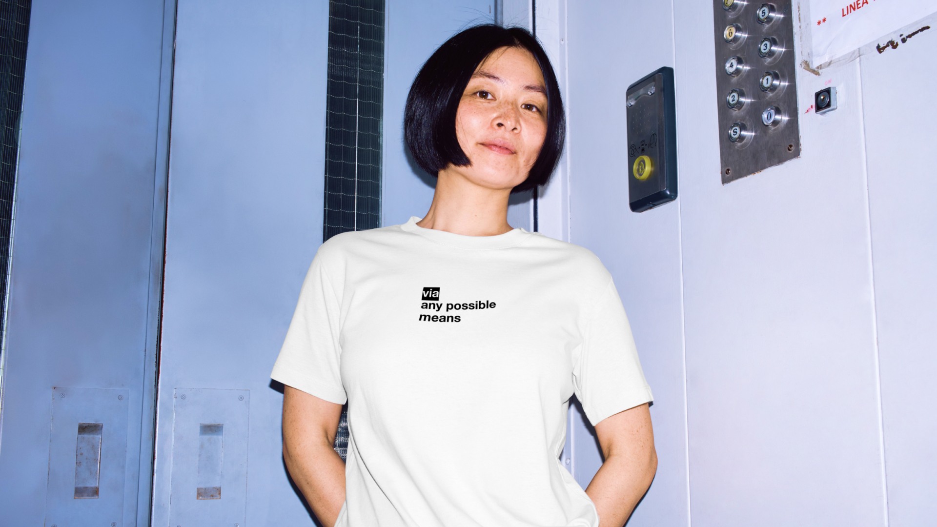 Woman with short black hair wearing a white shirt that says 'via any possible means' standing inside an elevator.