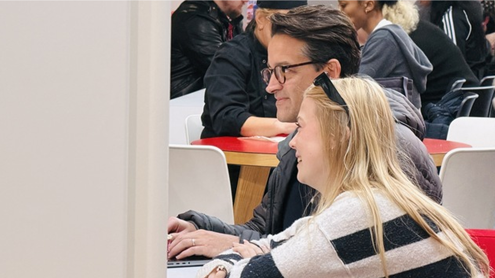 Smiling man and woman sitting side by side in a busy area, looking at a laptop screen.