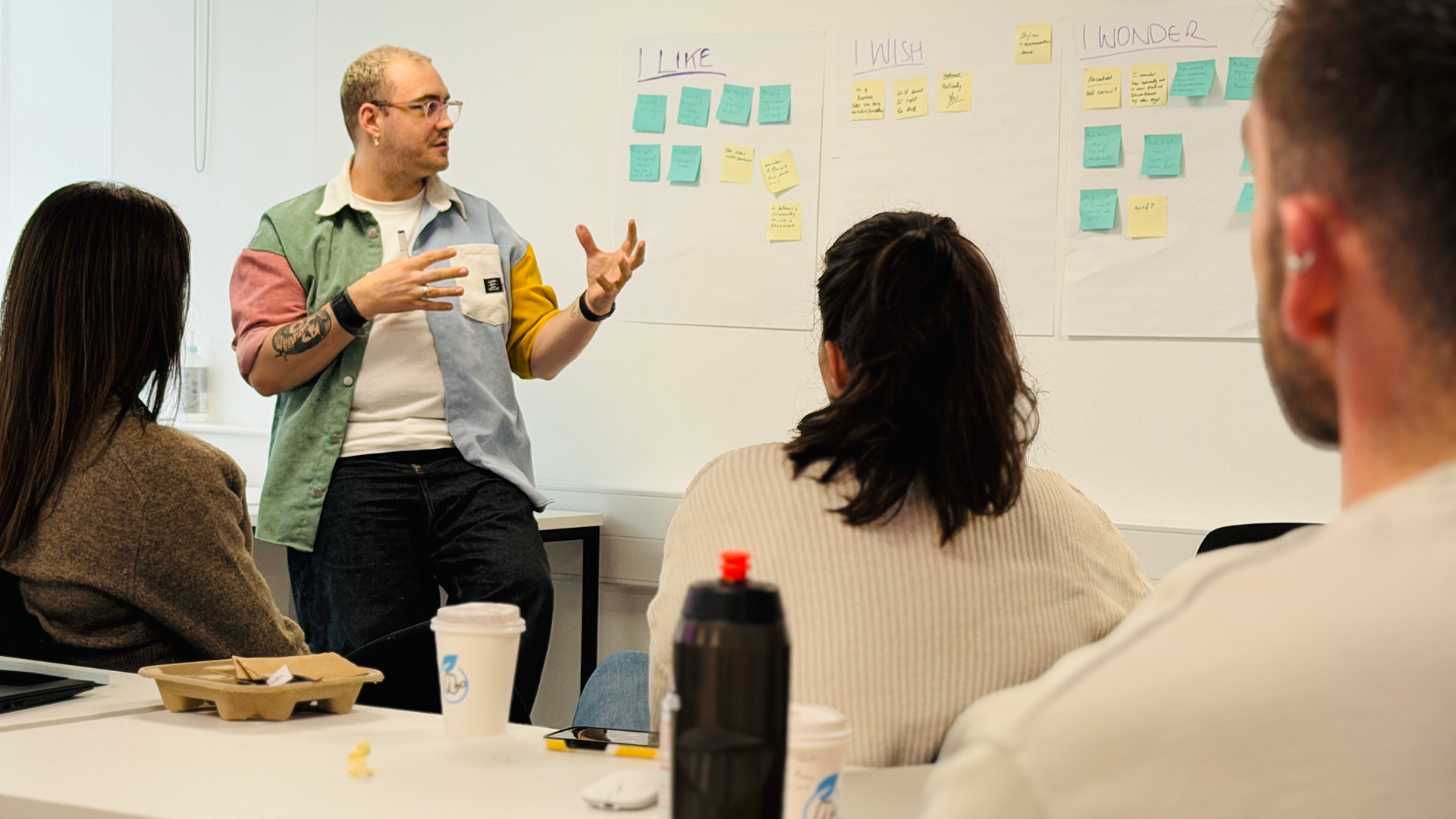 Man in a color-blocked shirt explaining ideas to a group of three people seated around a table with sticky notes on the wall labeled I LIKE, I WISH, and I WONDER.