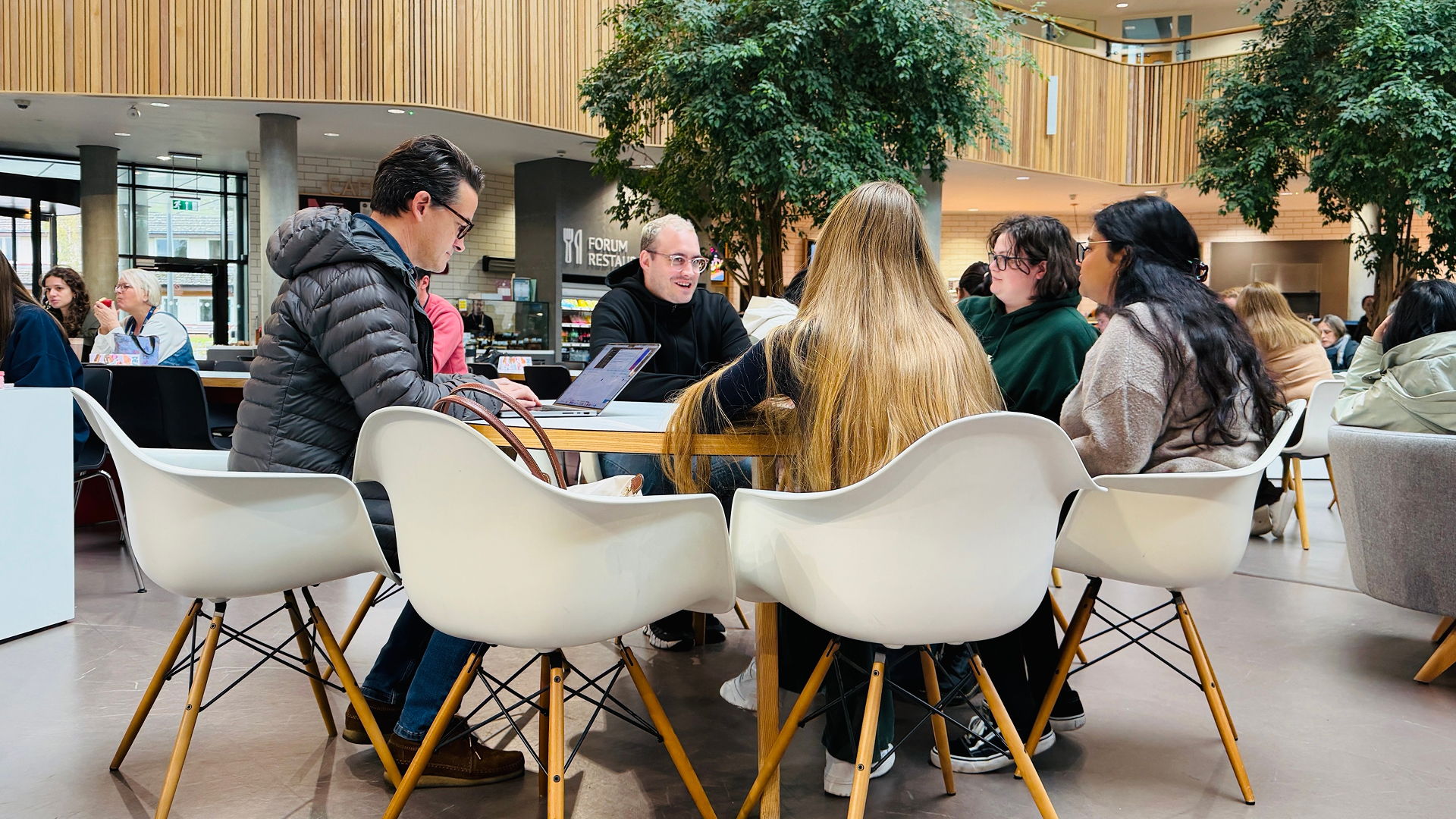 Group of young adults seated around a table in a modern indoor common area with plants and wooden accents, some using a laptop.