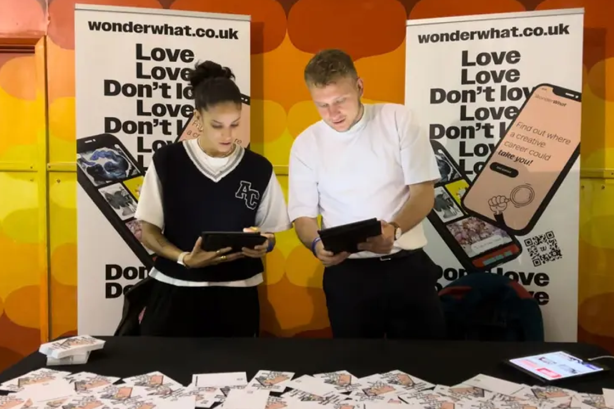 Two people standing behind a table with promotional materials, holding tablets, in front of 'wonderwhat.co.uk' banners.