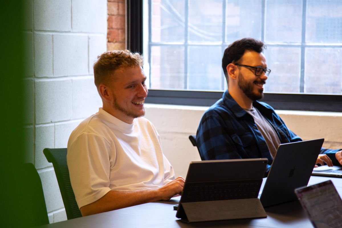 Two young men smiling and working on laptops at a table in a bright office with large windows.