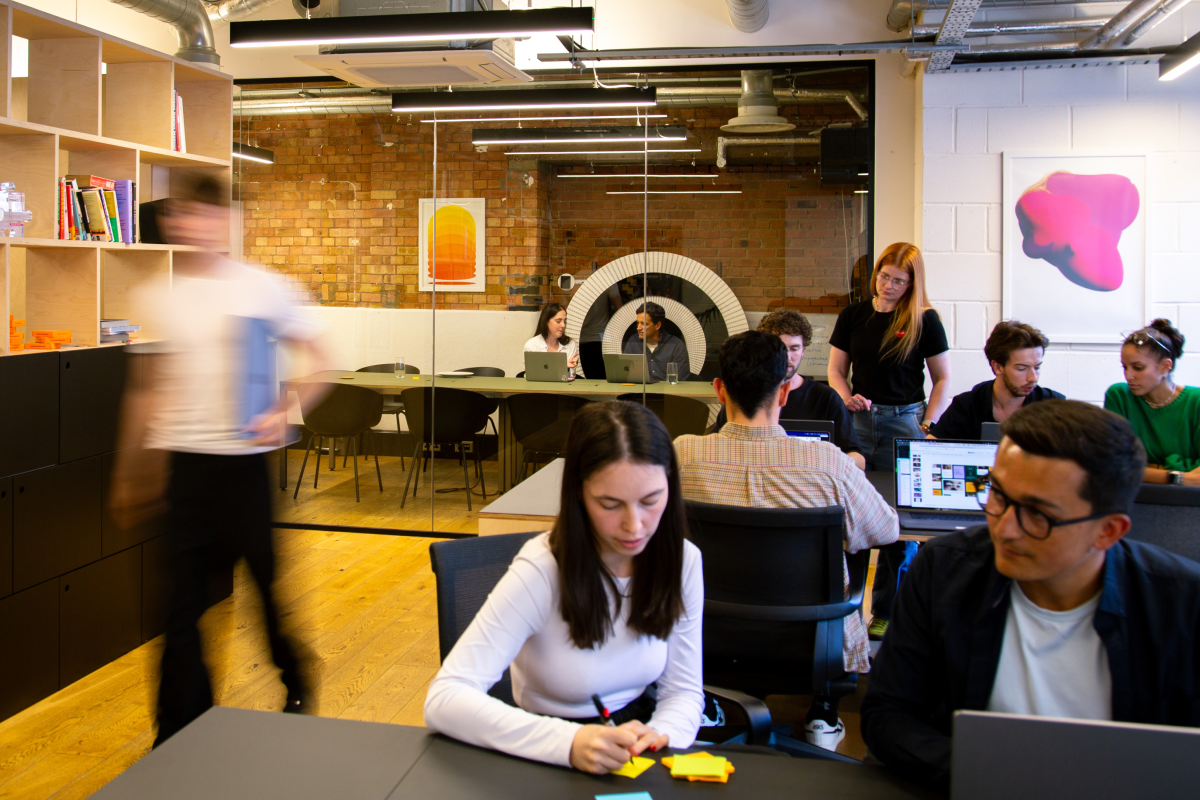 A modern office with multiple people working, including a woman writing on sticky notes and others using laptops or conversing.