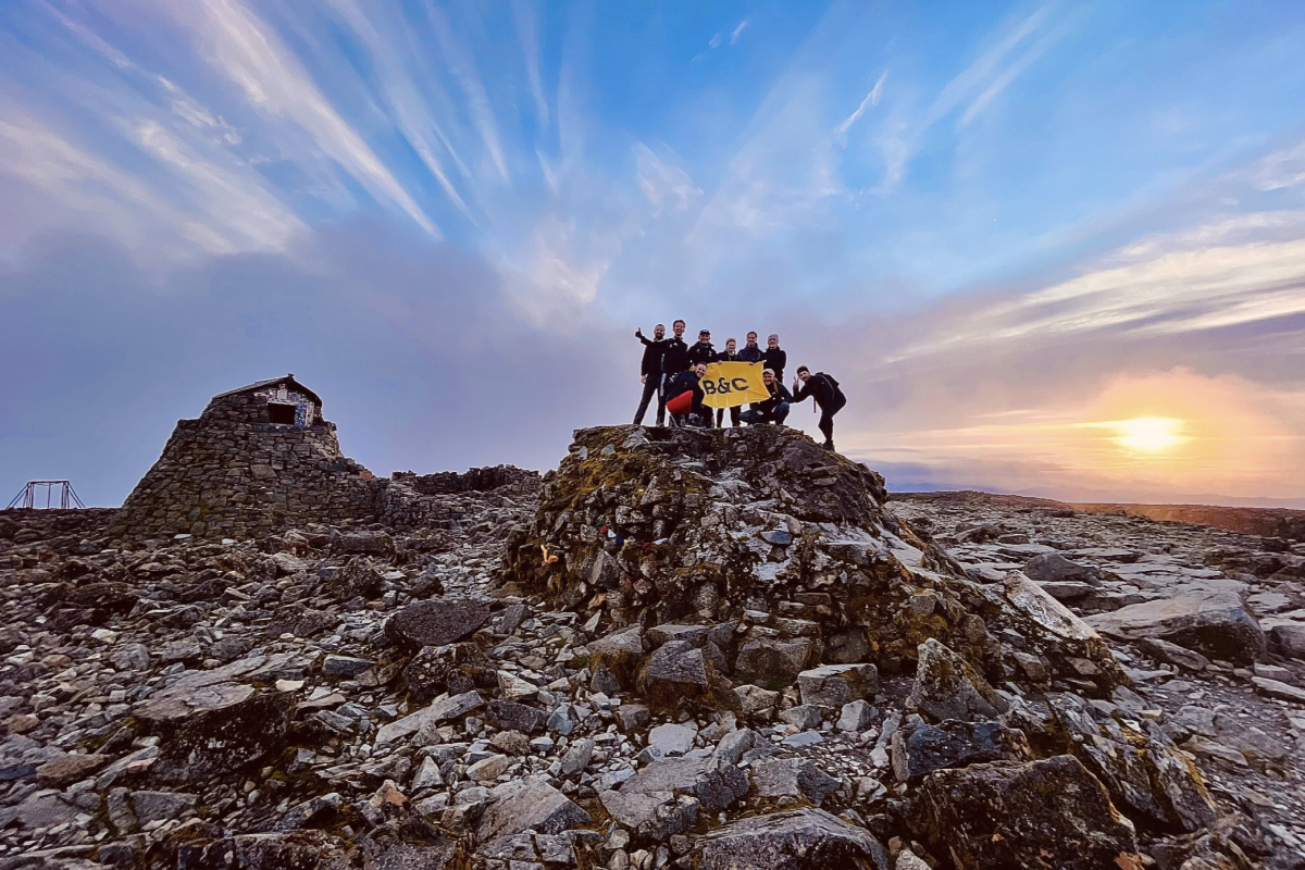 Group of people standing on the peak of Ben Nevis holding a yellow B&C banner at sunset with a stone structure in the background.