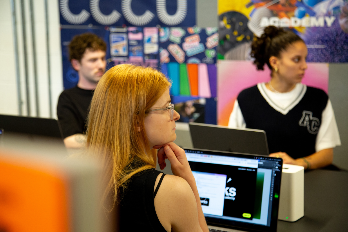 Three young adults in a colorful room work on laptops, with one red-haired woman in focus resting her chin on her hand.
