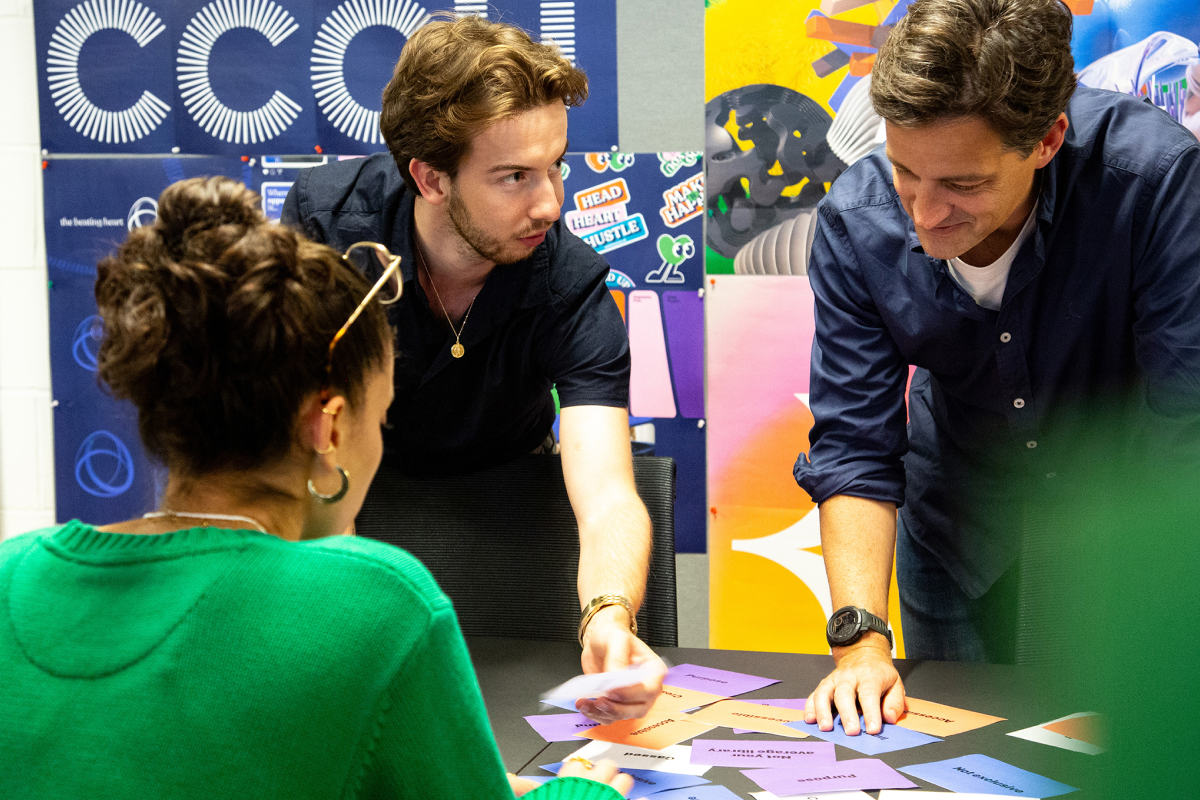 Three people in a discussion around a table with colorful paper cards spread out, with posters on the wall behind them.