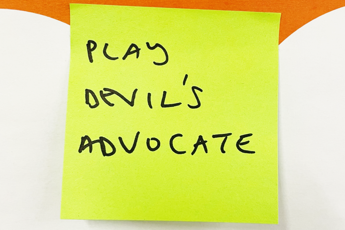 Yellow sticky note with handwritten text saying 'PLAY DEVIL'S ADVOCATE' on a white and orange background.