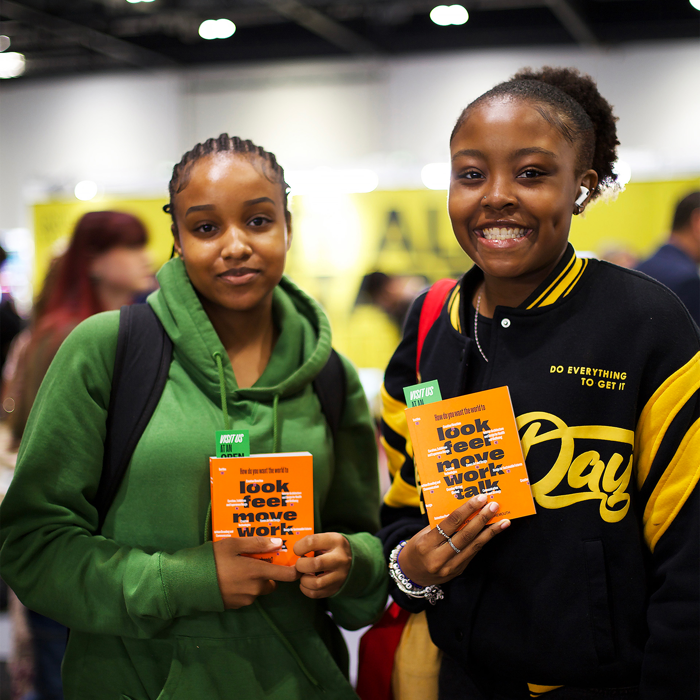 Two young women smiling and holding orange booklets titled 'look feel move work talk' at an event.