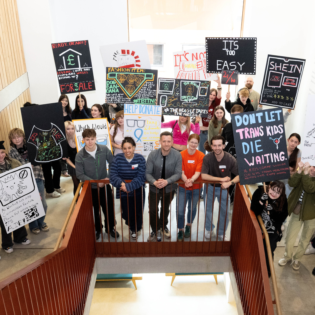 Group of diverse young people holding protest signs about social issues and standing behind a railing in a bright indoor space.