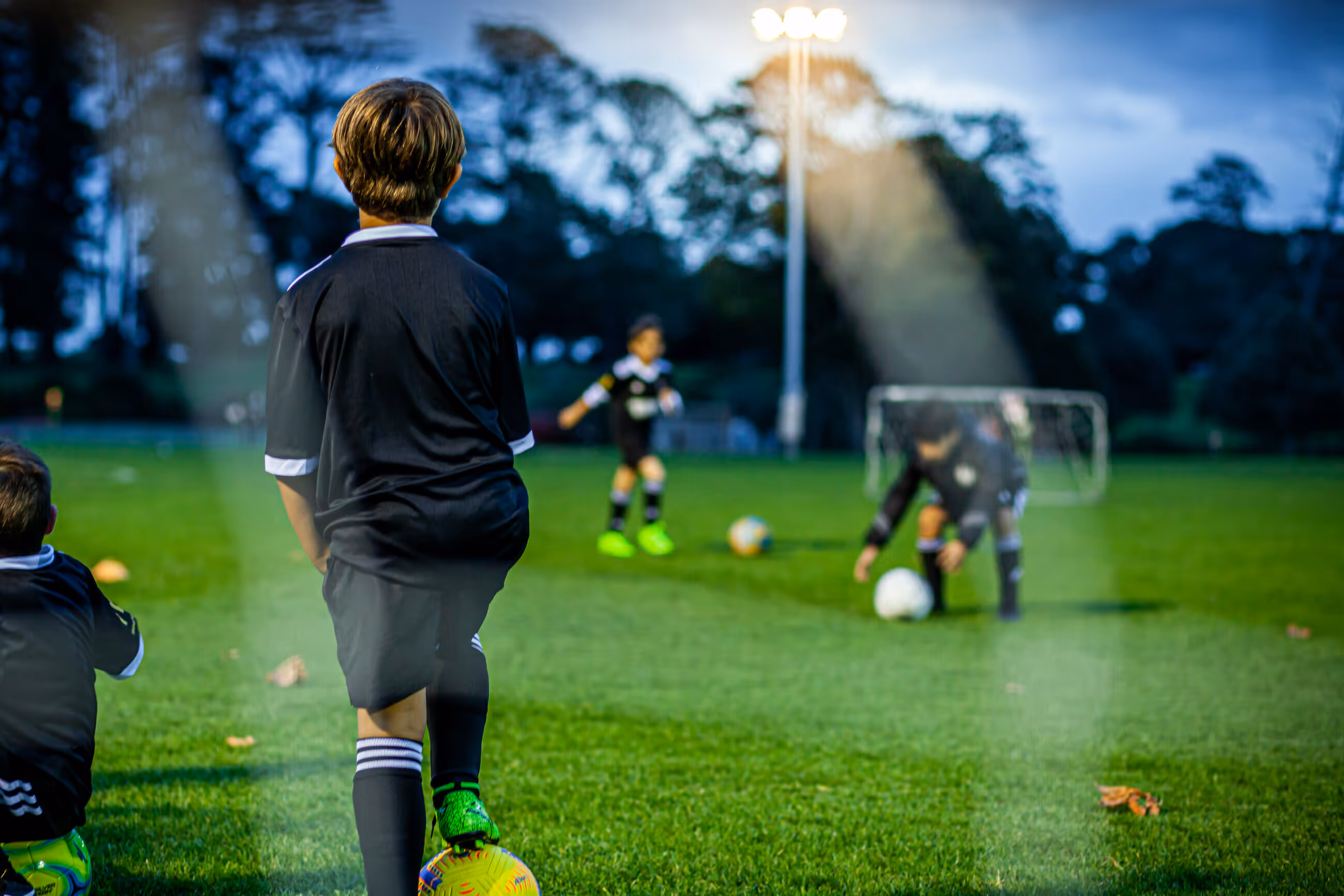 A young boy in a football kit seen from behind, standing with his foot on a ball, looking across a floodlit pitch towards other children practicing.