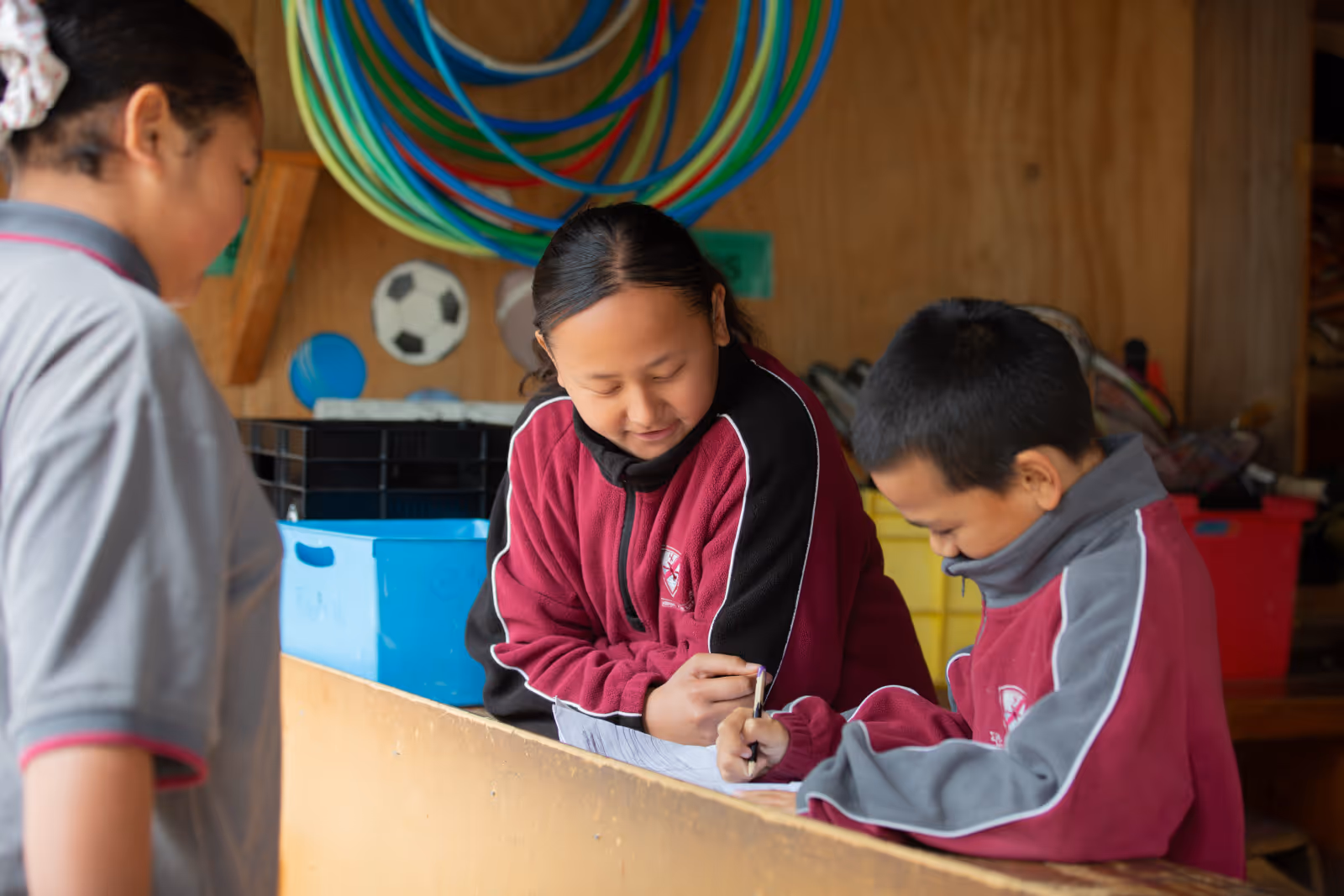 Three children in school uniforms engaged in writing and interacting in a PE shed