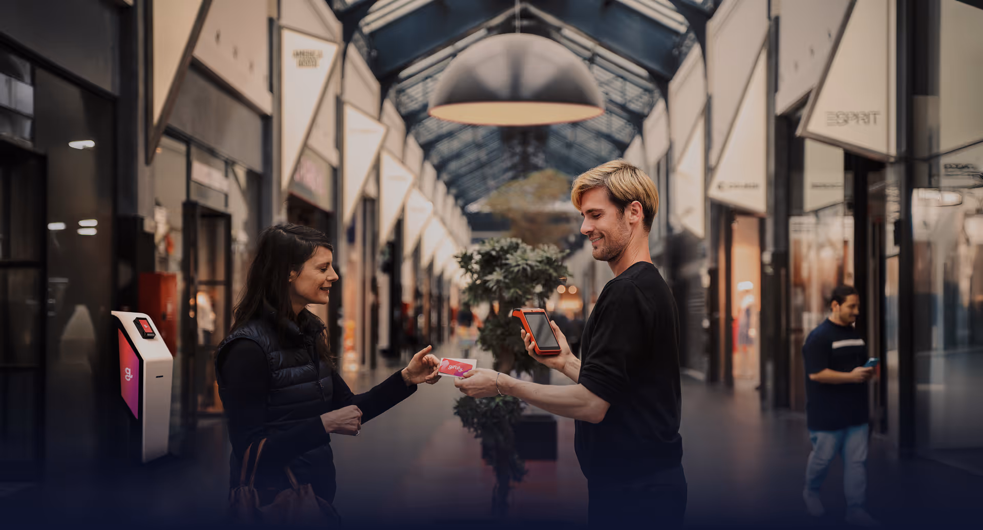 A Giftify representative hands a gift card to a woman in a shopping mall, with a Giftify kiosk visible in the background on the left.