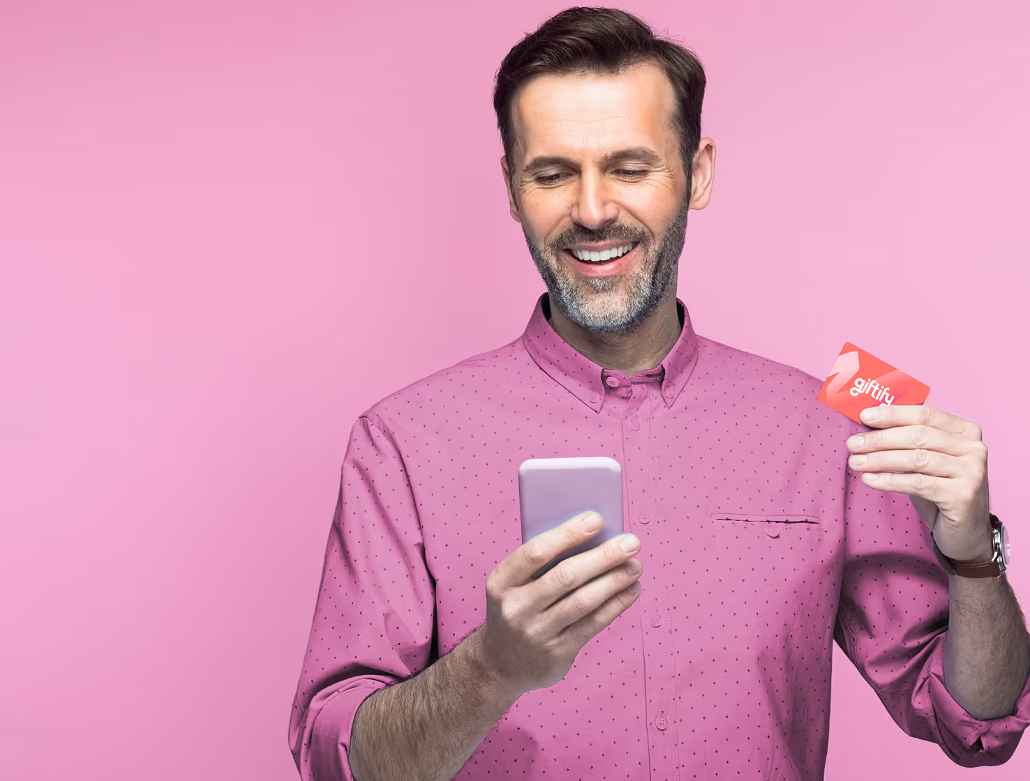A smiling man in a pink shirt holds a purple smartphone in one hand and a red Giftify gift card in the other. He looks down at his phone. The background is a solid pink.