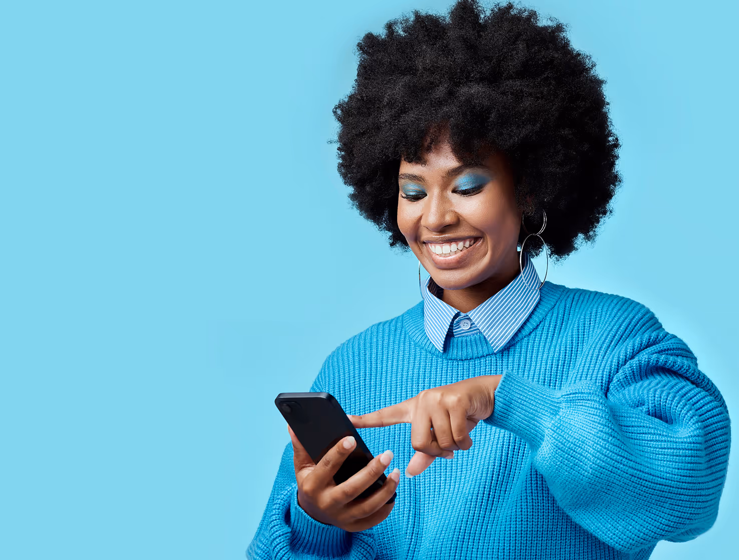 A happy woman with a large afro hairstyle and blue eyeshadow smiles as she uses her phone. She is wearing a blue sweater over a striped shirt. The background is a solid light blue.