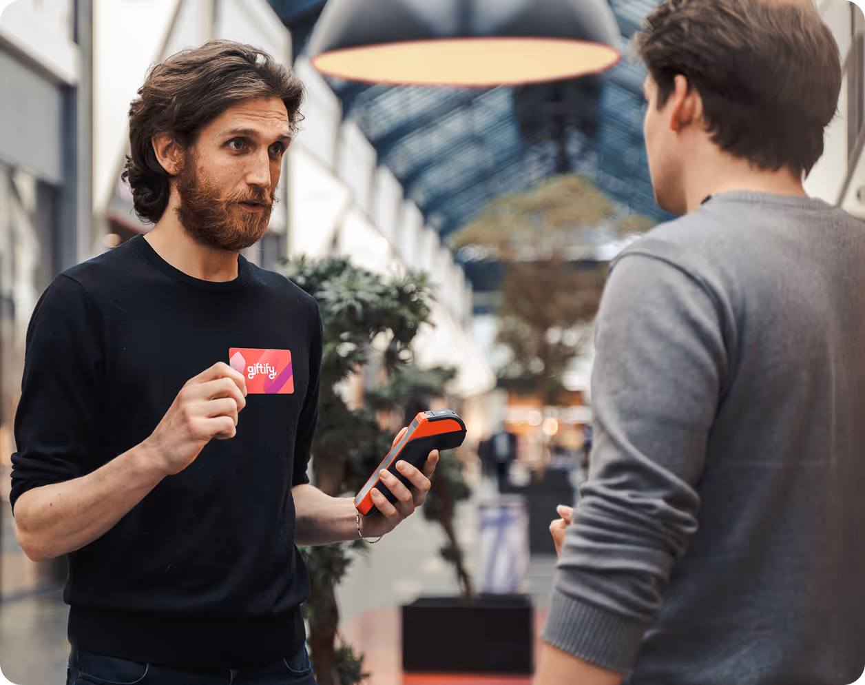 A man wearing a black shirt with the Giftify logo holds a pink Giftify gift card and a card reader, speaking to a customer in a modern retail space.