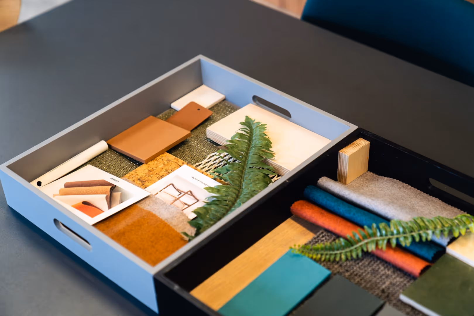 Interior design mood board featuring fabric swatches, wood samples, leather pieces, and printed photos, arranged neatly in a tray with greenery accents.