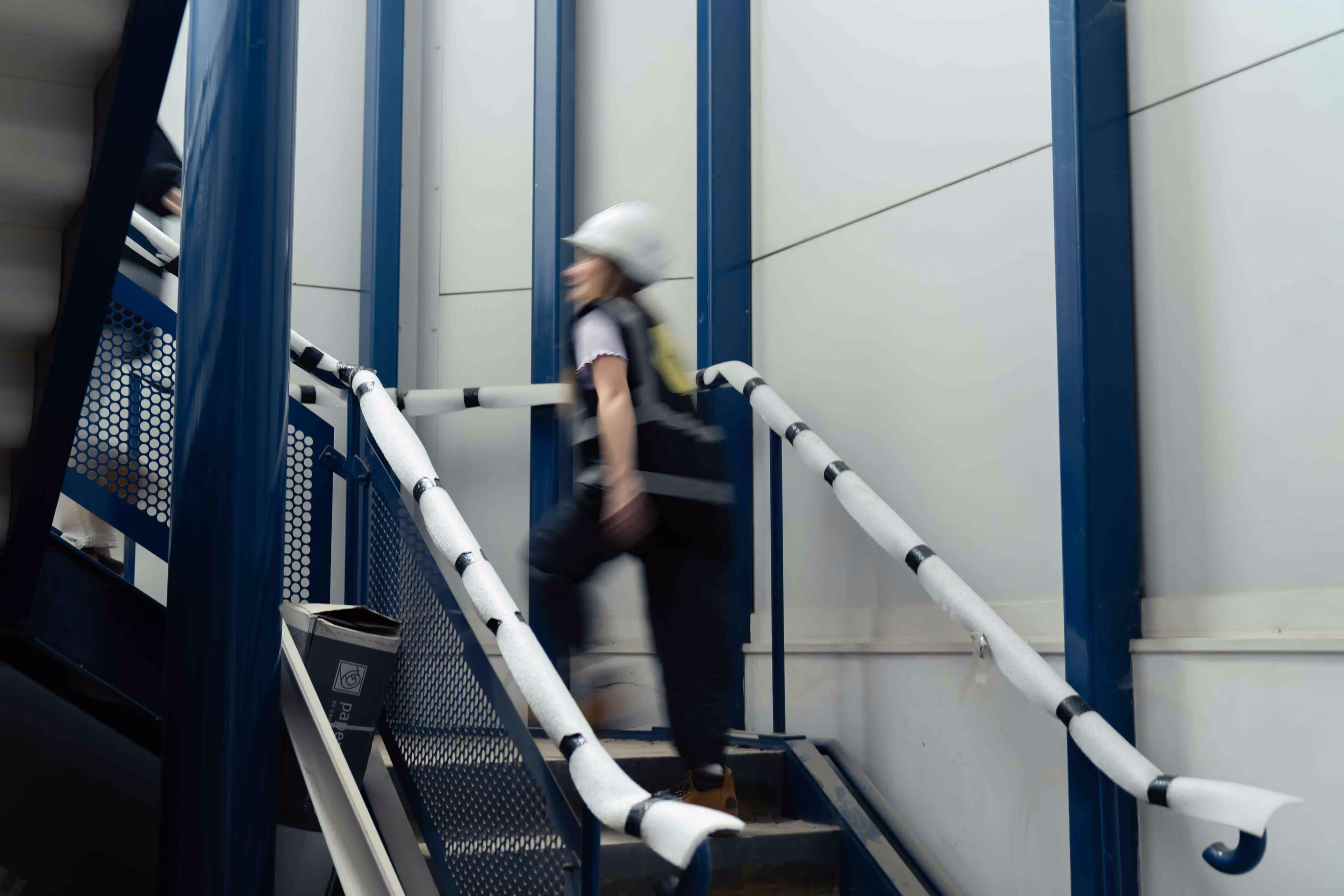 Person in safety gear and hard hat walking up a metal staircase inside an industrial or construction setting.