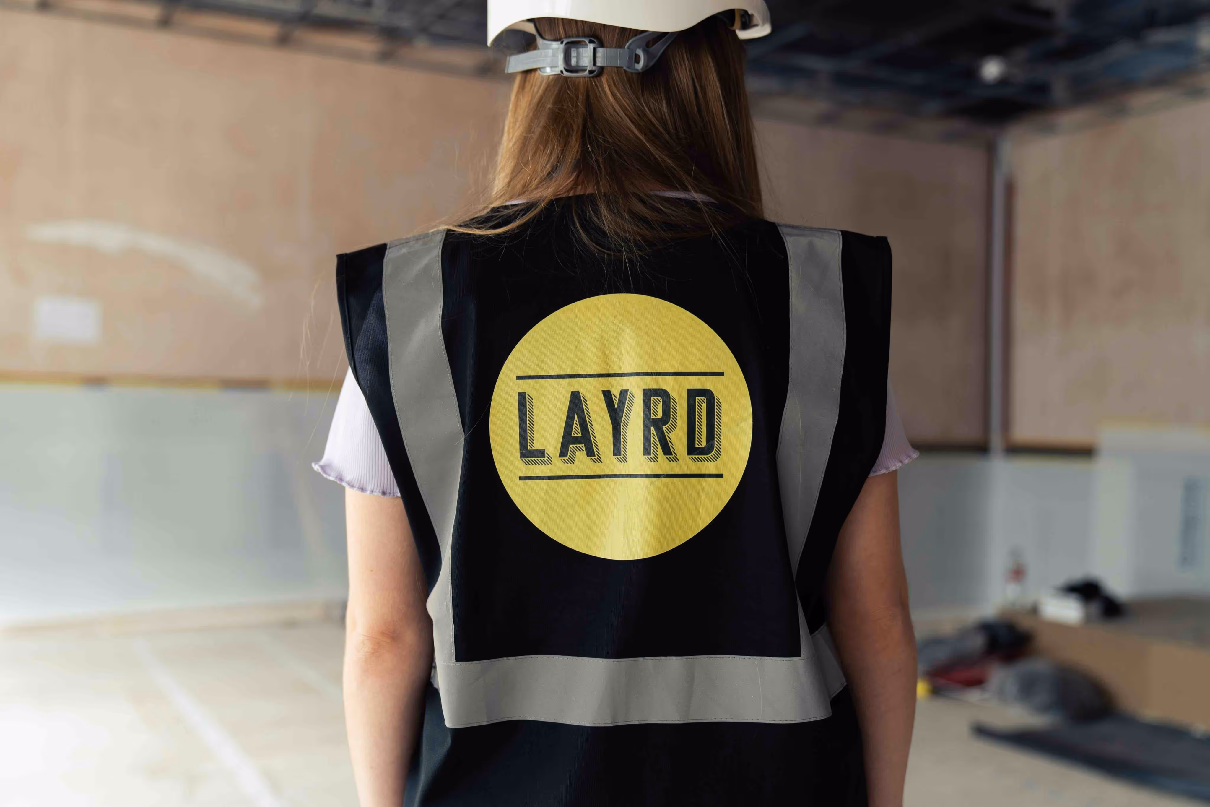 Person wearing a high-visibility vest and hard hat with 'LAYRD' logo on the back, standing in an unfinished interior space.