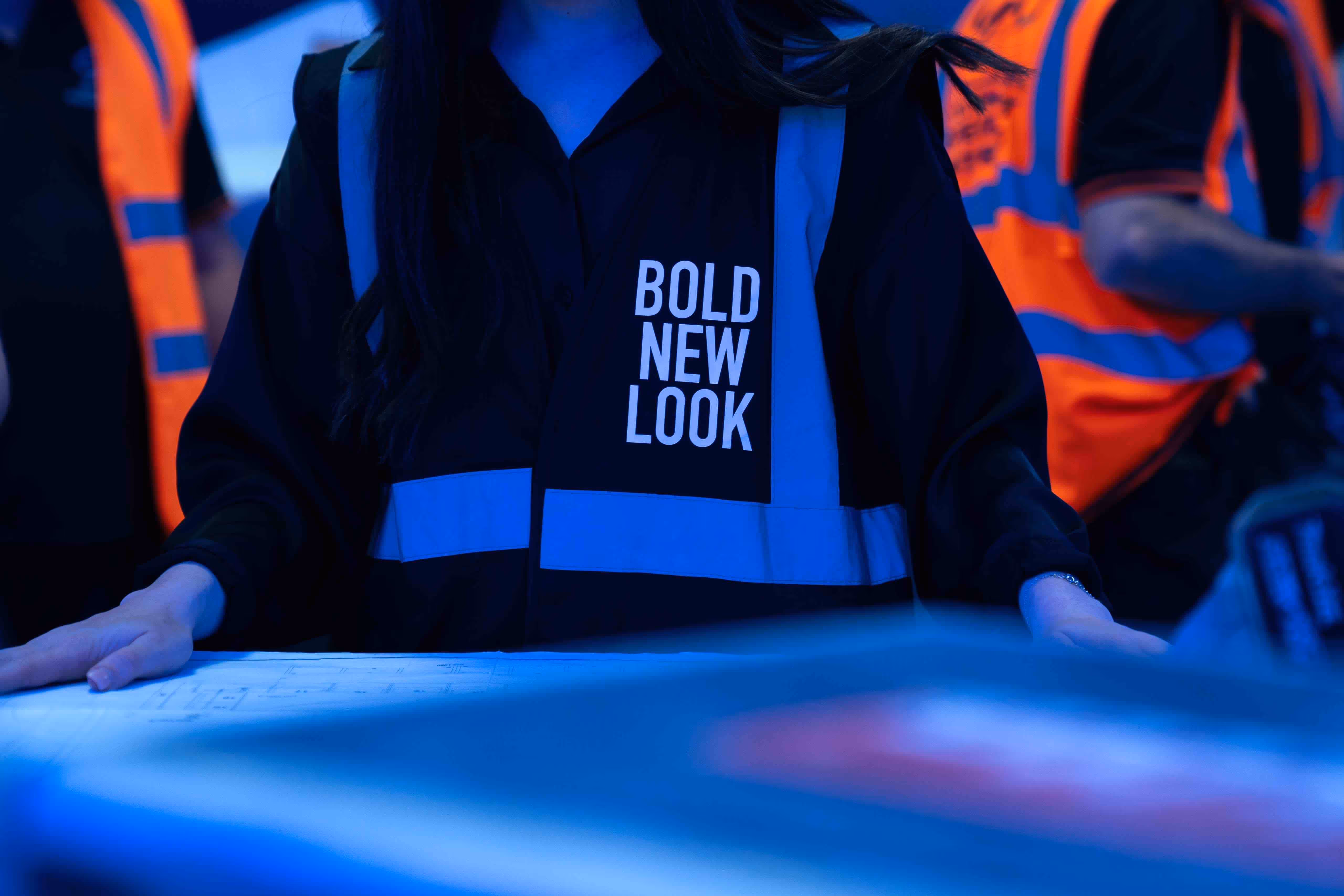 Person wearing a 'Bold New Look' vest reviewing plans at a construction or design site.