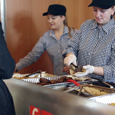 two women in gingham shirts, black hats and gloves plating tacos for customers at a Popup