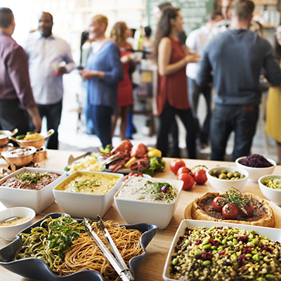 Table with beautiful catered foods in the foreground, while an office party goes on behind it.