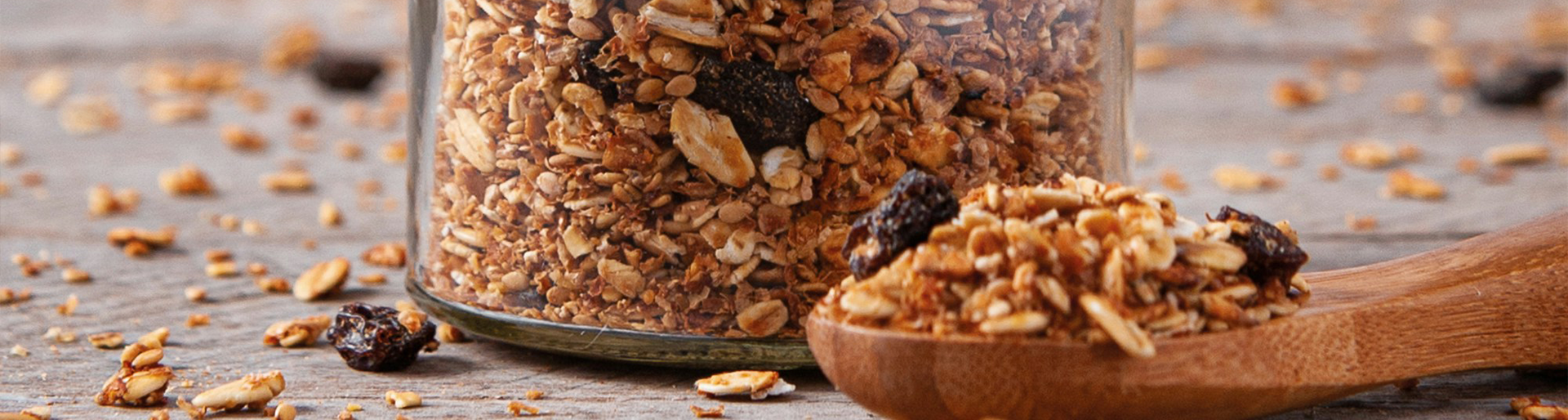 spoon of granola in the foreground with the bottom of a glass jar filled with granola behind.g