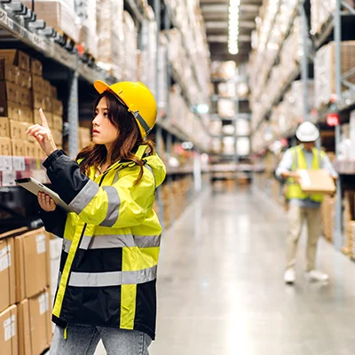 A woman in a hi-vis jacket stands in a warehouse aisle with a clipboard looking at the stacked boxes.