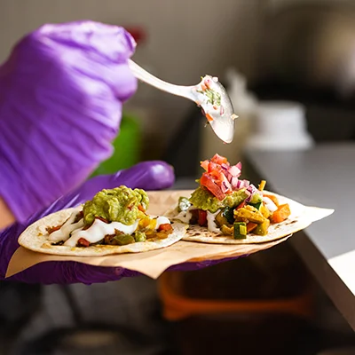 hands in purple gloves spooning pico de gallo onto two tacos.