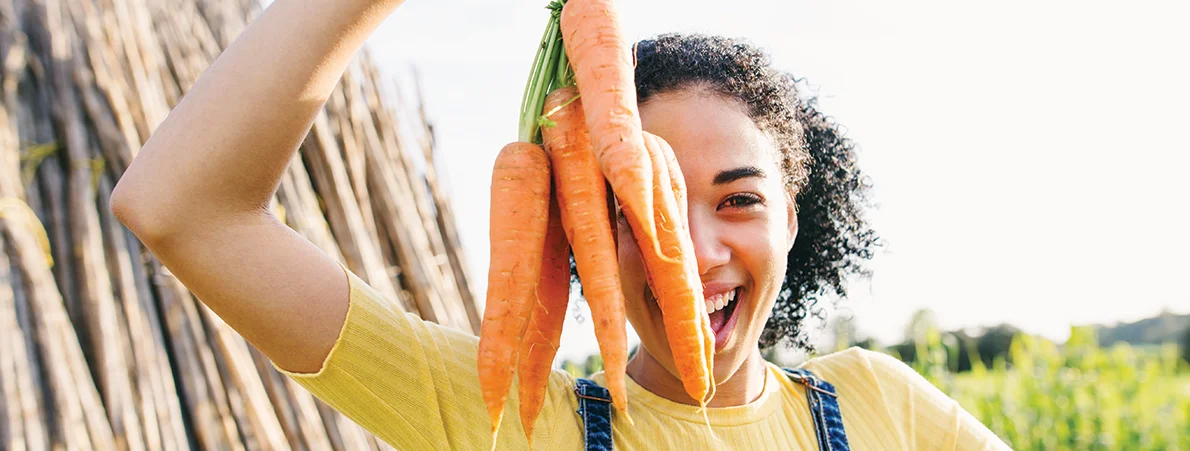 young woman holding a bunch of carrots up in front of her face on a farm.