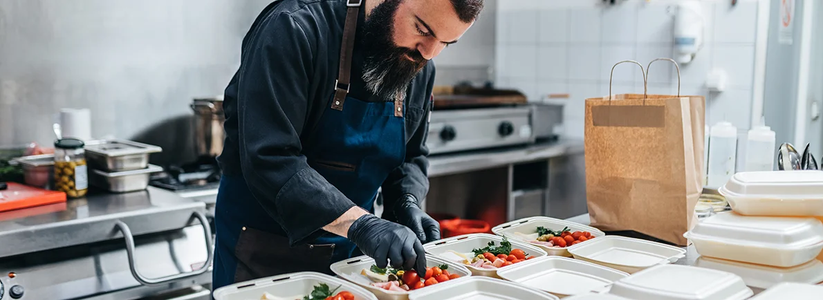 Male chef filling to-go orders in a restaurant kitchen with fresh seasonal ingredients.